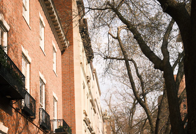 Brick apartment building with black iron balconies along a sunlit, open sidewalk