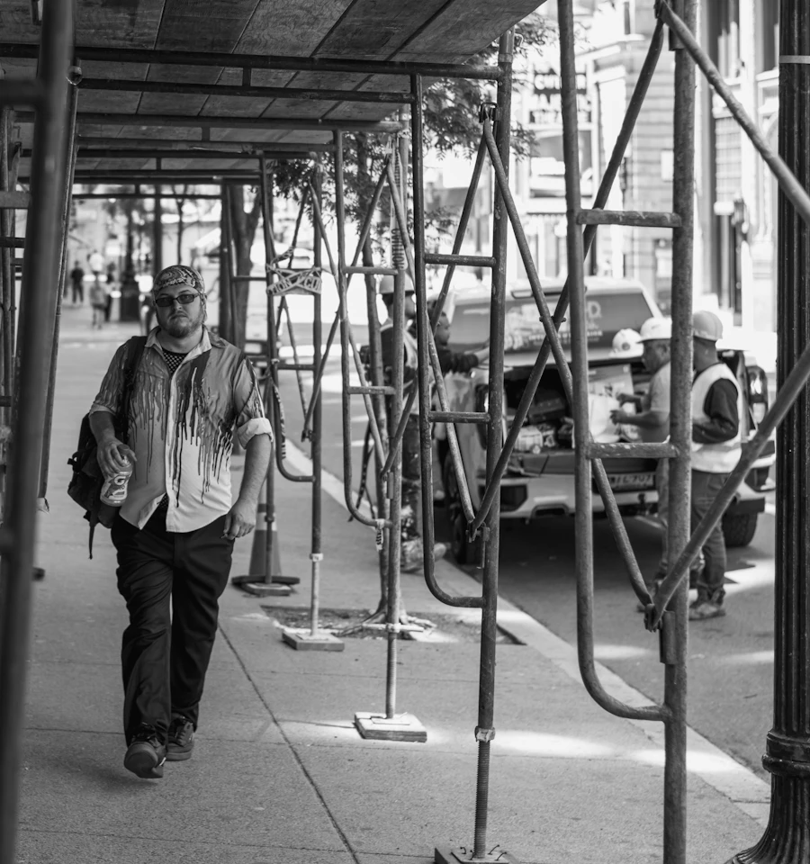 Pedestrian walking through a covered sidewalk under metal scaffolding on a city street
